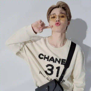 穿著米色 Chanel 衛衣的人，在白色牆前做出俏皮手勢。