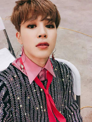 身穿閃亮黑色外套、粉紅色襯衫和紅色領帶的年輕人坐在戶外 — Jimin GIF 壁紙