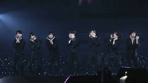 七位男性K-pop表演者在黑暗舞台上，伴隨藍色燈光和閃爍地板演唱。