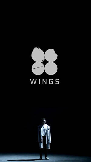 一個身穿白色外套的人，在黑暗的舞台上站在一個發光的「WINGS」標誌下方。