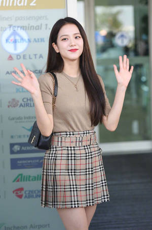 年輕女子在機場航站樓穿著格子裙和米色上衣向鏡頭揮手。