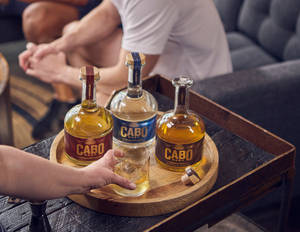 手將龍舌蘭酒倒入木托盤上的玻璃杯，旁邊有三支Cabo Wabo酒瓶。
