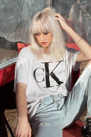 年輕女性留著金色頭髮，穿著Calvin Klein Jeans T恤和破洞牛仔褲，坐在紅色椅子上。