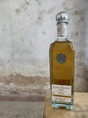 一瓶Casa Noble Reposado龍舌蘭酒放在木頭上，背景是混凝土牆。 — 3:4 壁紙