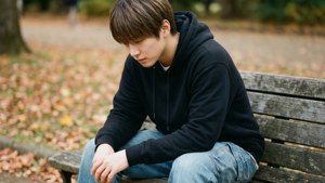 穿著黑色連帽衫的年輕人獨自坐在公園木長椅上，周圍是落葉