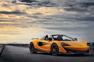 日落時分，戲劇性的雲層下，橙色McLaren跑車停在路上。 — MacBook 壁紙