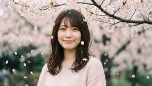 一位微笑的女性站在春天公園裡落下的櫻花下。