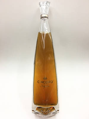 一個透明玻璃瓶Cincoro Tequila，裝有金色液體，背景為白色。