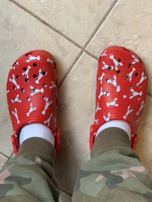 米色瓷磚地板上的紅色Crocs鞋，帶有白色龍蝦圖案，搭配迷彩褲和白色襪子。