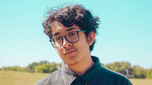 戴眼鏡的年輕男子站在陽光明媚的田野中，背景有樹木。