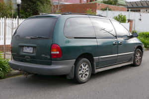 深綠色Chrysler Voyager小型貨車停泊喺住宅區街道，有白色籬笆同磚牆。 — MacBook Air M2 壁紙