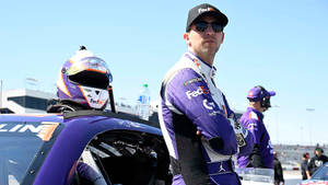 身穿紫色制服的NASCAR車手站在賽道上的賽車旁邊。