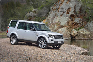 銀色 Land Rover Discovery 停泊在礫石河岸上，有岩石懸崖和樹木。