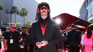 身穿黑色西裝和紅色背心的男子，戴著太陽眼鏡和耳機在紅地毯上擺姿勢。