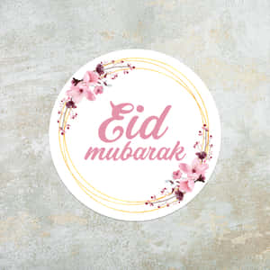 圓形 Eid mubarak 問候貼紙，帶有粉紅色花朵和金色線條，位於紋理灰色表面上。