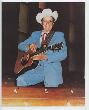 一位穿著藍色西裝和牛仔帽的老年男子在舞台上彈奏原聲吉他。