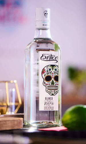 Exotico Blanco 龍舌蘭酒瓶，帶有糖骷髏標籤，旁邊有青檸和酒吧用具。