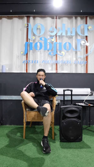 男子坐在藤椅上，在綠色草地上拿著咪高峰，旁邊有揚聲器。