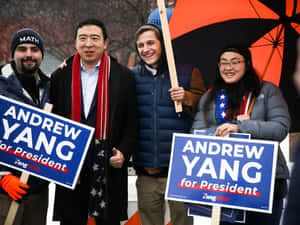 四個人穿著冬季裝備，在橙色雨傘下舉著Andrew Yang競選總統標語牌。 — 政治 壁紙