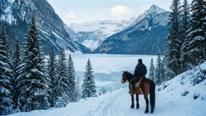 人在冰凍湖泊旁的雪徑上騎馬，周圍有白雪覆蓋的松樹和山脈