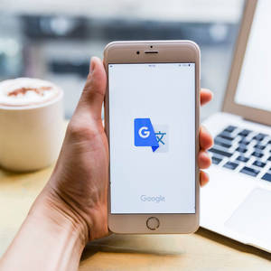 手拿著手機，Google翻譯應用程式打開，旁邊有手提電腦和咖啡。