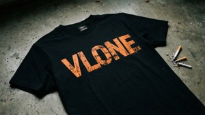 黑色Vlone T恤，橙色文字，在混凝土地板上，旁邊有煙頭