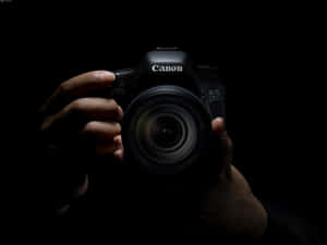 在黑暗的攝影棚環境中，雙手拿著一部黑色的Canon DSLR相機。 — 攝影師 壁紙