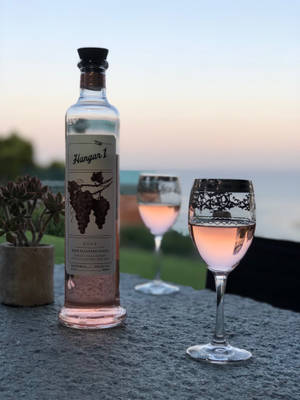 兩個酒杯和一瓶玫瑰伏特加放在石檯上，享有日落海景。 — 玫瑰金 壁紙