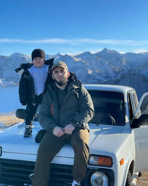 男子和小孩坐在白色汽車引擎蓋上，背景是積雪山景。