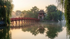 紅色橋樑和寶塔倒映在霧氣瀰漫的水中，周圍環繞著垂柳，黎明時分