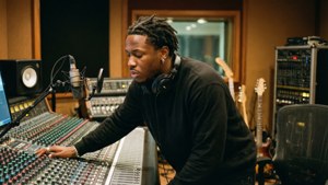 一位音樂家在錄音室中調整音頻設備，背景有吉他。