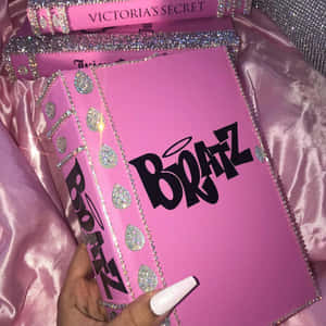 手拿著一本帶有閃粉和水晶的粉紅色 Bratz 書，放在緞面布料上。