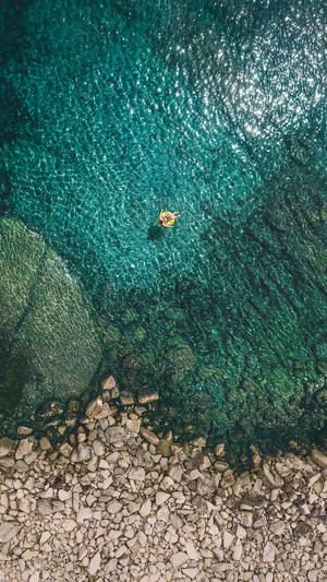 一個人喺岩石海岸附近嘅清澈碧綠海水中，漂浮喺一個黃色充氣浮床上。 — 沙灘 壁紙