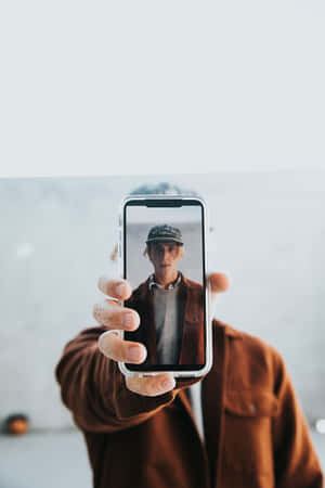 人拿著手機顯示自拍照，穿著棕色外套和帽子