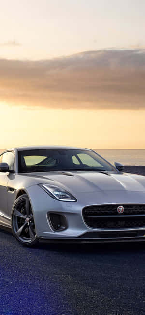 銀色跑車停在夕陽下的海灘上，背景是海洋 — iPhone 15 Plus 壁紙