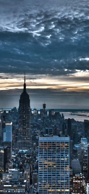 暮色中的紐約市天際線，帝國大廈在烏雲和水面上亮起燈光。 — iPhone 15 Pro 壁紙