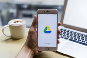 手裡拿著手機，打開了 Google Drive 應用程式，旁邊有咖啡和手提電腦。