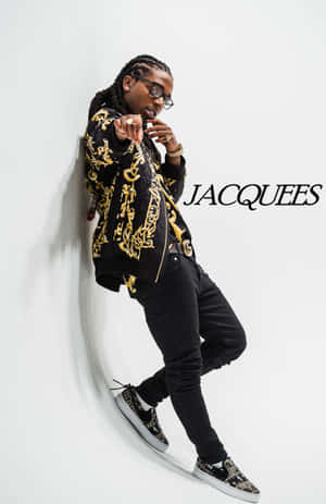 身穿金色圖案外套和運動鞋的男士靠著白牆，帶有 'JACQUEES' 文字。