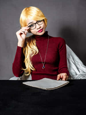 金髮戴眼鏡的女性在書桌前調整眼鏡，桌上放著筆記本。 — 哈莉·奎茵 壁紙