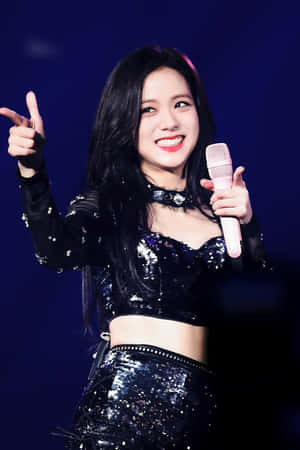K-pop表演者穿著閃亮服裝在舞台上微笑並用麥克風指向觀眾。