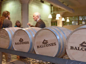 帶有 Balcones 標誌的木製威士忌桶，釀酒廠內人們在交談。 — 政治 壁紙