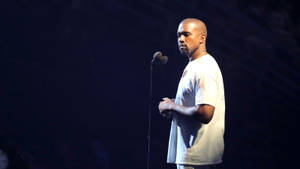 Kanye West 在黑暗舞台上於聚光燈下表演，穿著白色T恤。