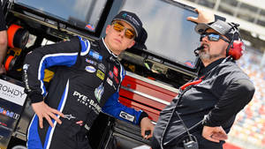 兩名身穿賽車裝備的NASCAR車隊成員站在維修區的賽車旁。
