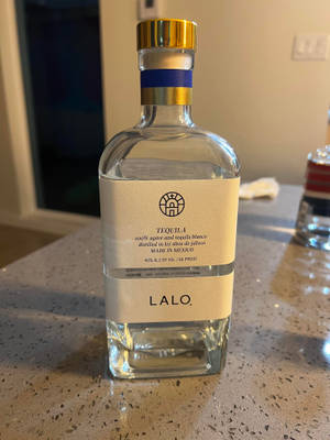 一個透明嘅龍舌蘭酒樽，金色樽蓋同忌廉色標籤，喺有斑點嘅枱面上。