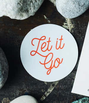 白色圓形貼紙，橙色草書文字寫著 Let it Go，被灰色石頭包圍。 — 滾石樂隊 壁紙