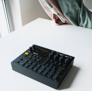 窗邊白色桌上的黑色 Digitakt 音樂序列器，帶有垂掛布料。