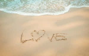沙灘上的沙中畫有心形的「LOVE」字樣，海浪湧入。 — 2880x1800 壁紙
