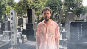 身穿粉紅色襯衫的男子站在日本墓地中，周圍有石頭紀念碑和綠樹。