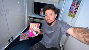 男人在細小房間裡拿著漫畫書，旁邊有電視和海報。