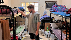 男子在雜亂的宿舍房間裡整理衣服，房間裡有雙層床。